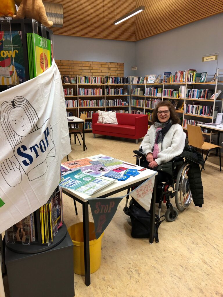 StoP-Aktive neben Infostand in einer Bibliothek.