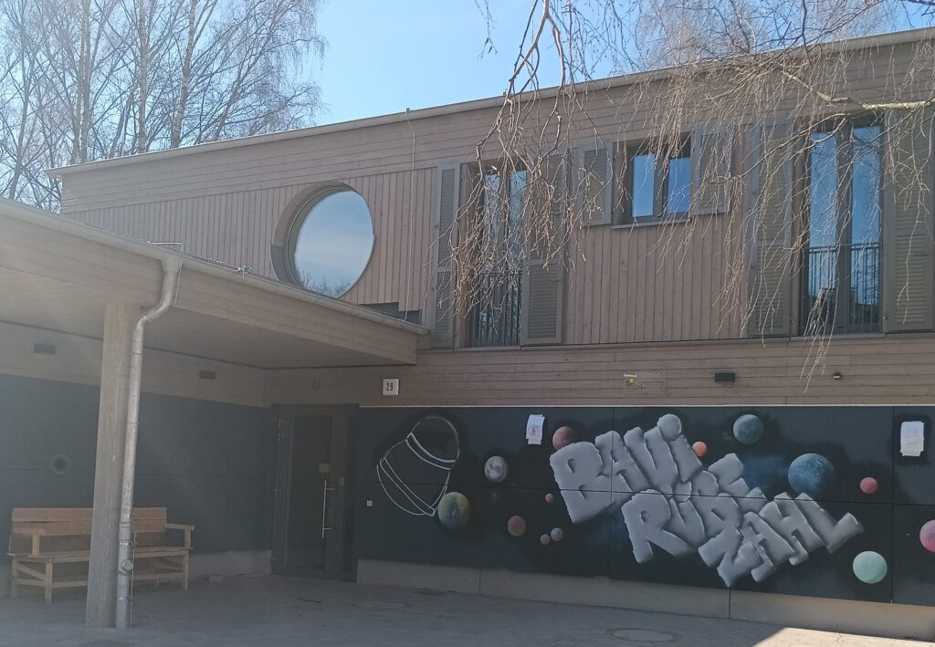 Vorderansicht vom Haus des Bauspielplatz Rübezahl mit dem Schriftzug BAUI RÜBEZAHL und der Eingangstür.