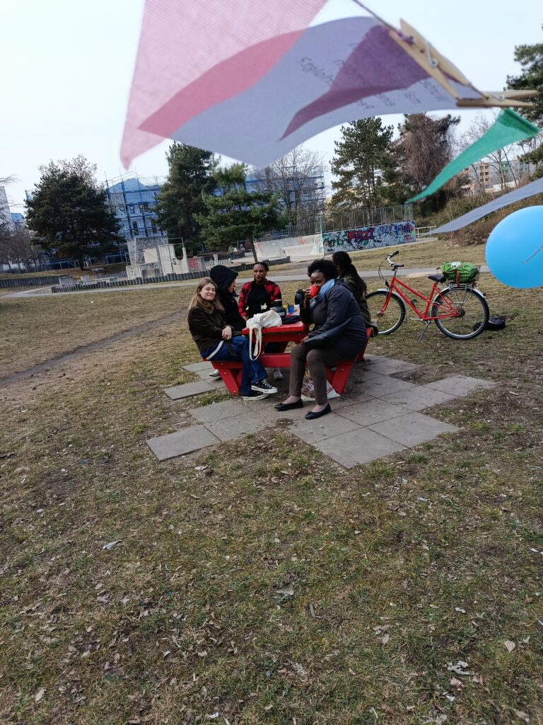 Gruppe an Frauen sitzen draußen an einer roten Bank im Grünen. Rechts neben sieht man angeschnitten im Winde wehen einen Luftballon an einer bunten Wimpelkette.